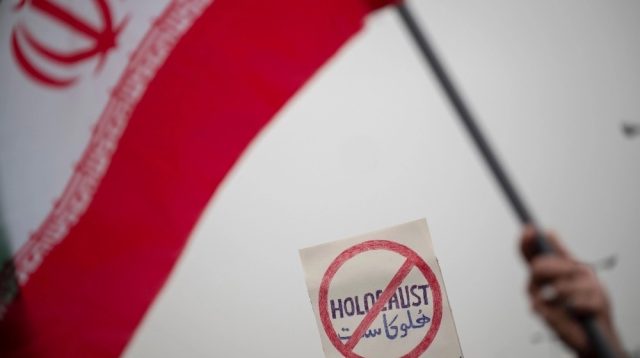 Seorang jamaah mengibarkan bendera Iran di depan plakat anti-Holocaust saat menghadiri protes atas pembakaran Al-Qu'ran setelah upacara Salat Jumat Teheran di masjid Agung Imam Khomeini, 27 Januari 2023. (Foto oleh Morteza Nikoubaz)